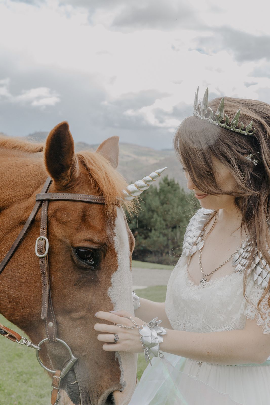 Bride wearing crown gently touches chestnut horse decorated as unicorn in outdoor setting.