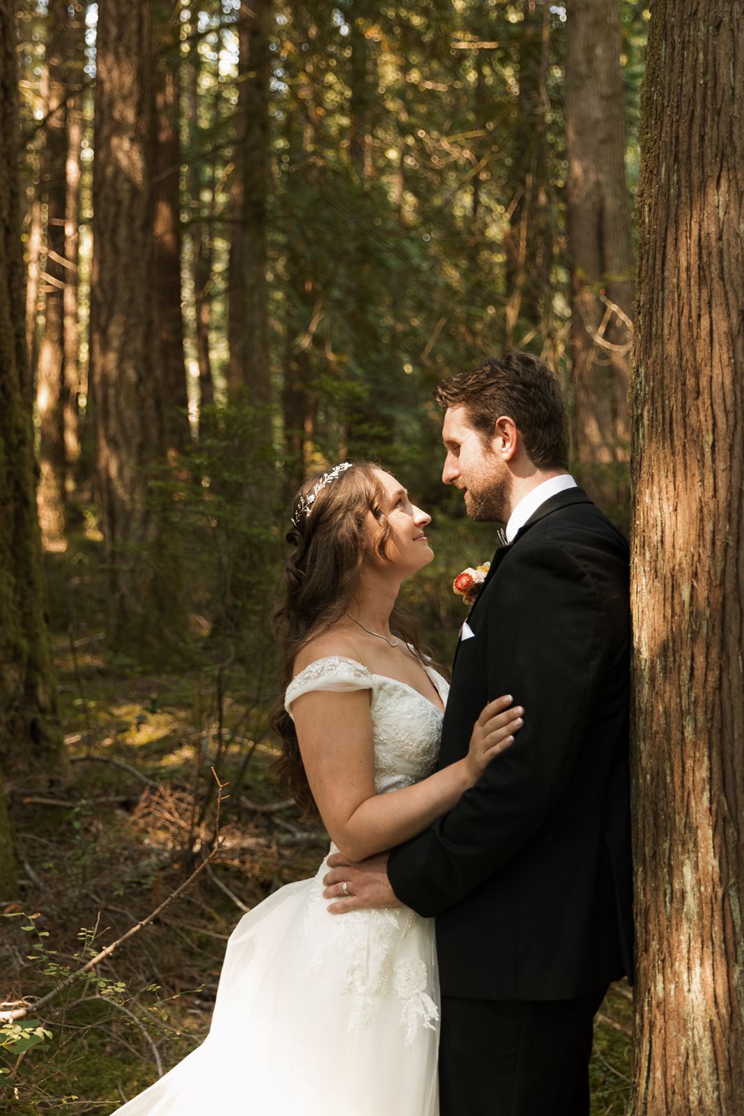 Bride and groom share a romantic moment leaning against a tree in a sunlit forest.