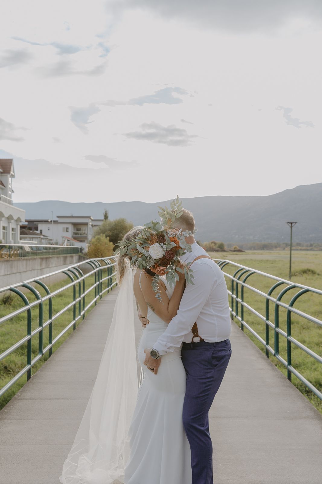 Bride and groom kissing behind a large floral bouquet on a bridge with mountain scenery on their wedding day.