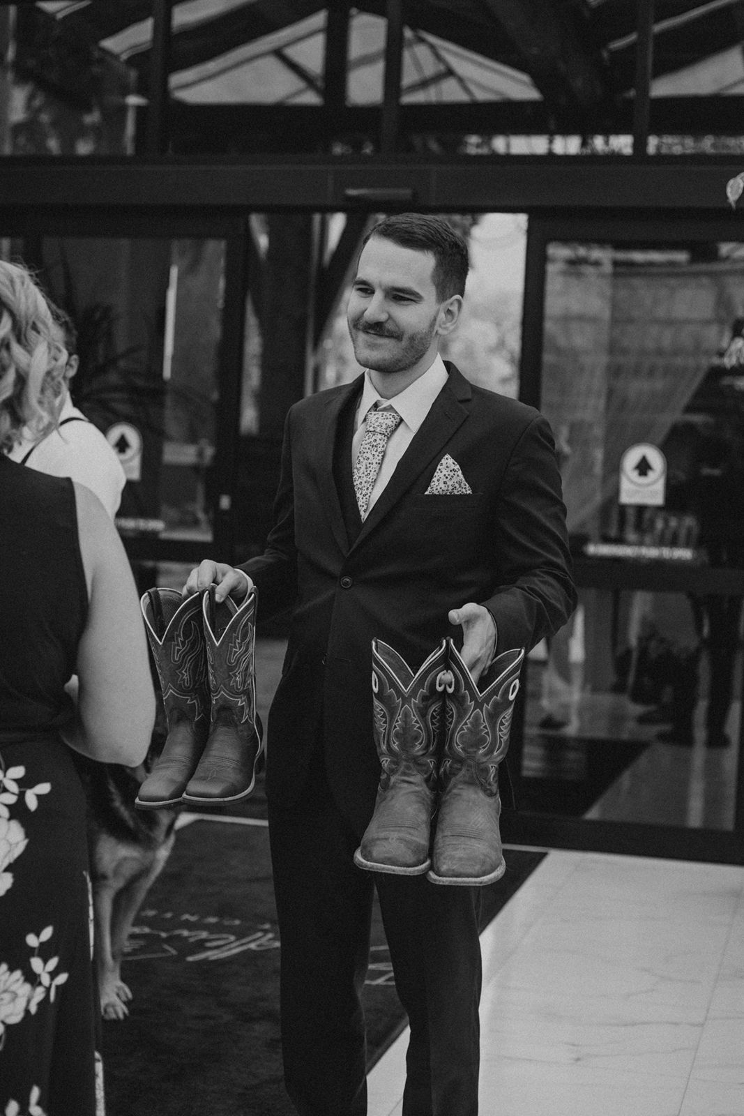 Black and white photo of smiling groom holding two pairs of cowboy boots at wedding reception.