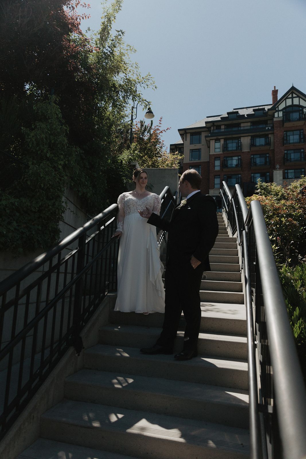 Bride and groom share a romantic moment on outdoor stairs with lush greenery and a brick hotel in the background.