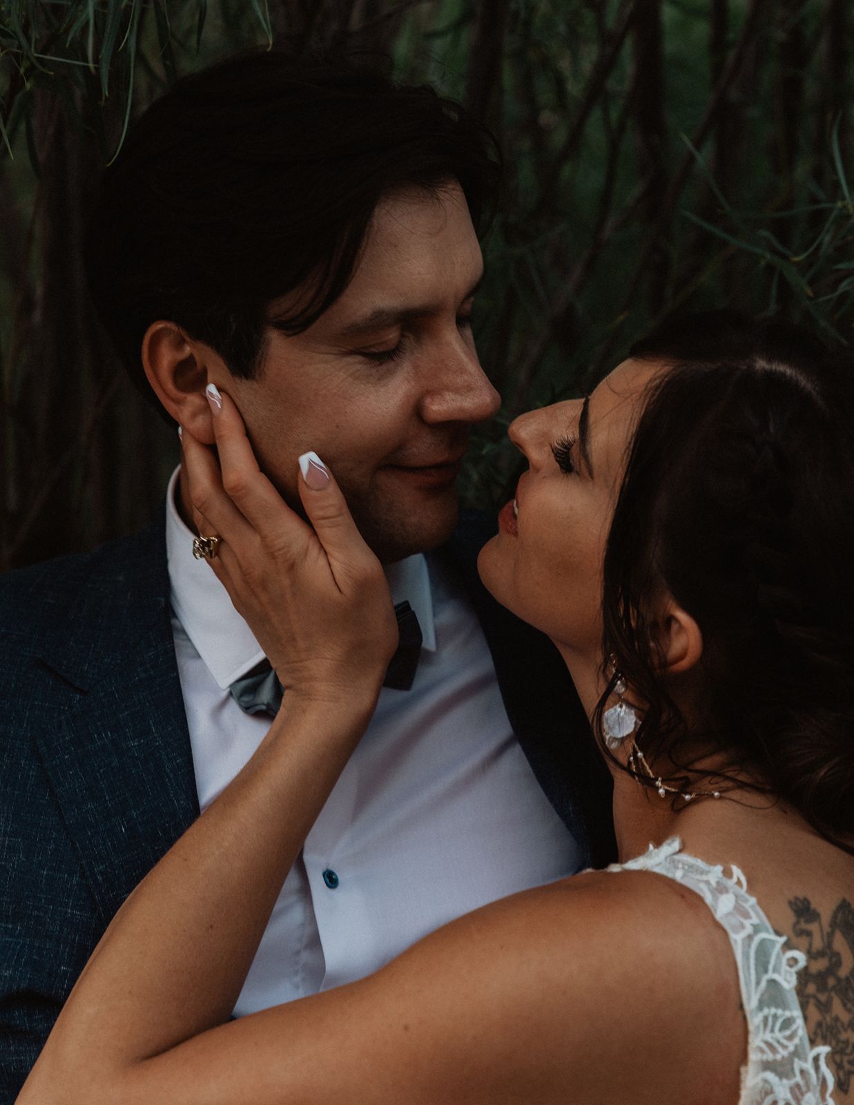 Couple sharing romantic almost-kiss moment surrounded by lush green foliage outdoors.