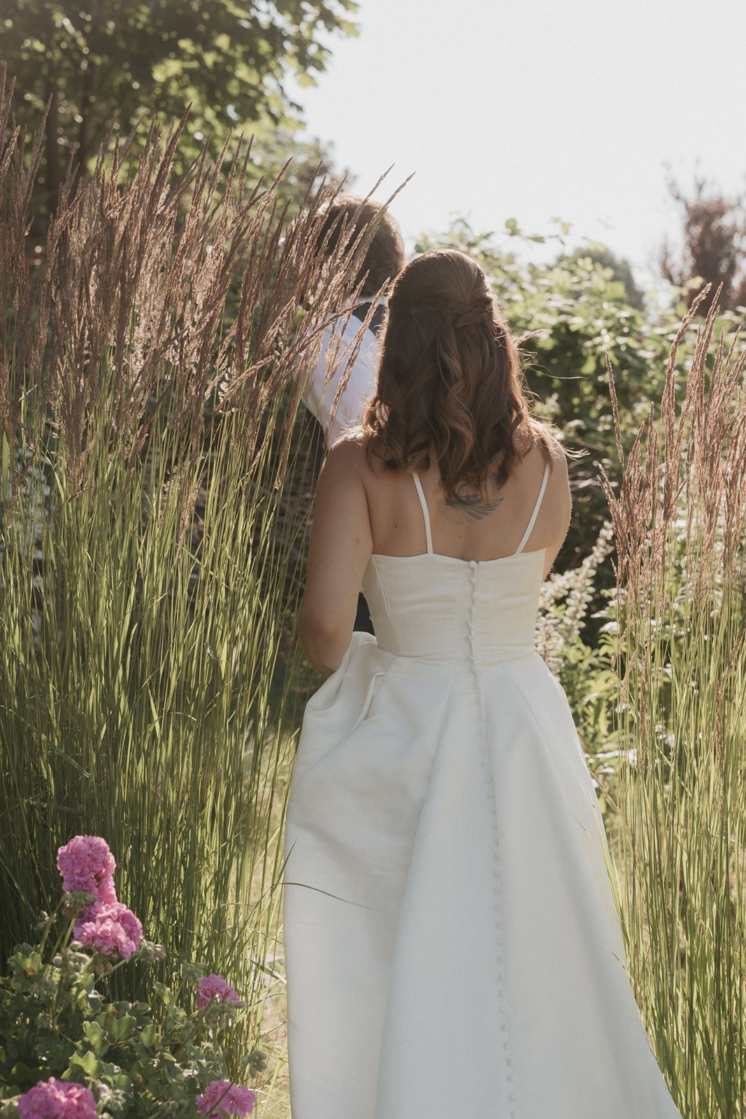 A bride in a white spaghetti-strap gown with button back walks through a sunlit garden path.