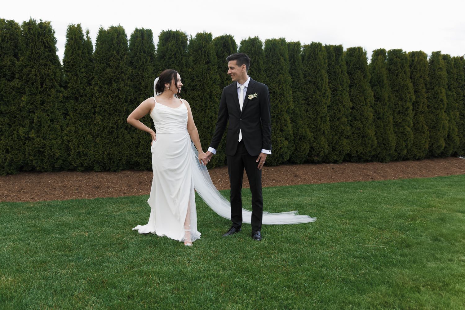 A bride and groom holding hands on a green lawn, smiling at each other, with tall hedges in the background.