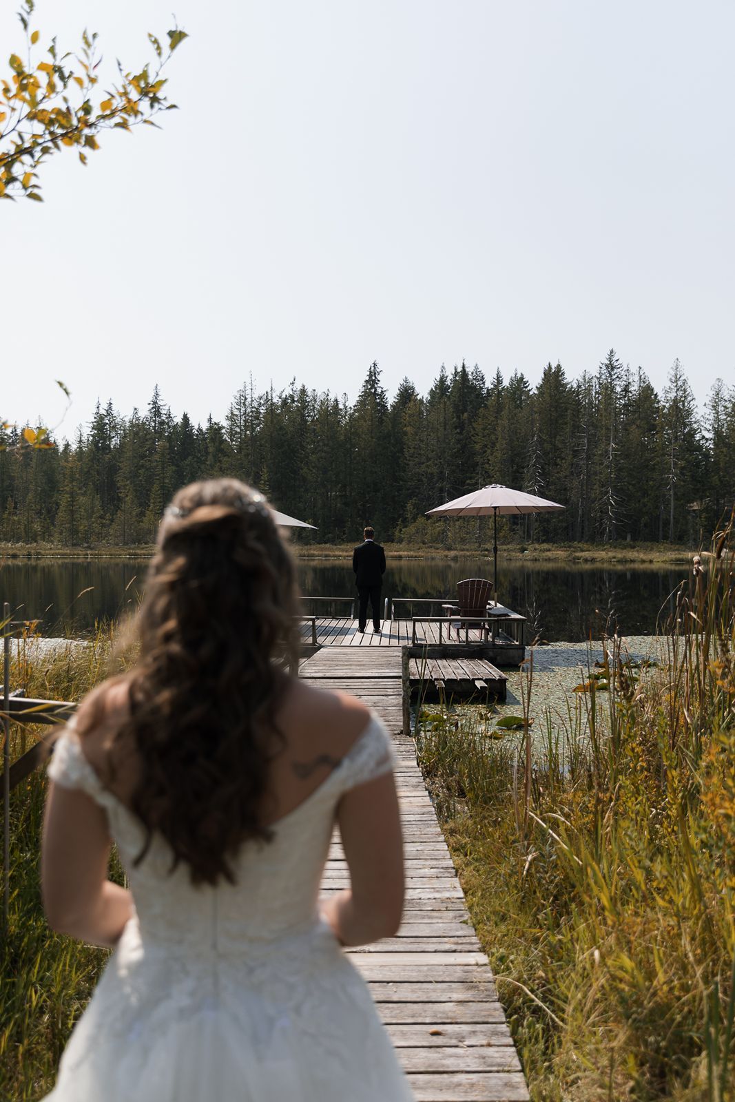 Bride in white gown walks toward groom standing on a wooden dock by a serene forest lake.
