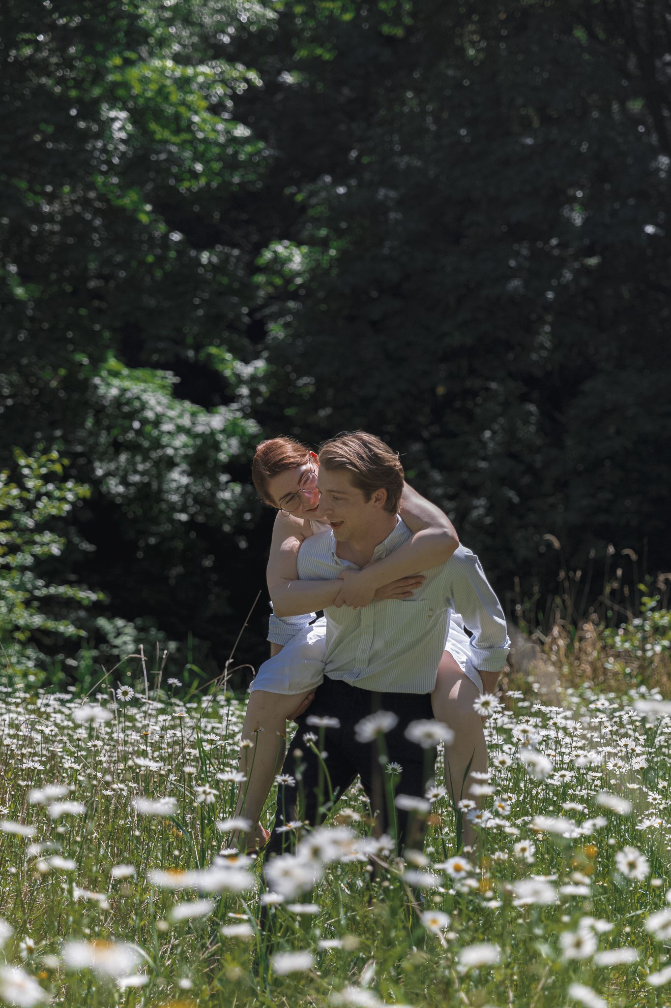 Groom giving bride a piggyback ride through a sunny wildflower meadow surrounded by lush green trees.