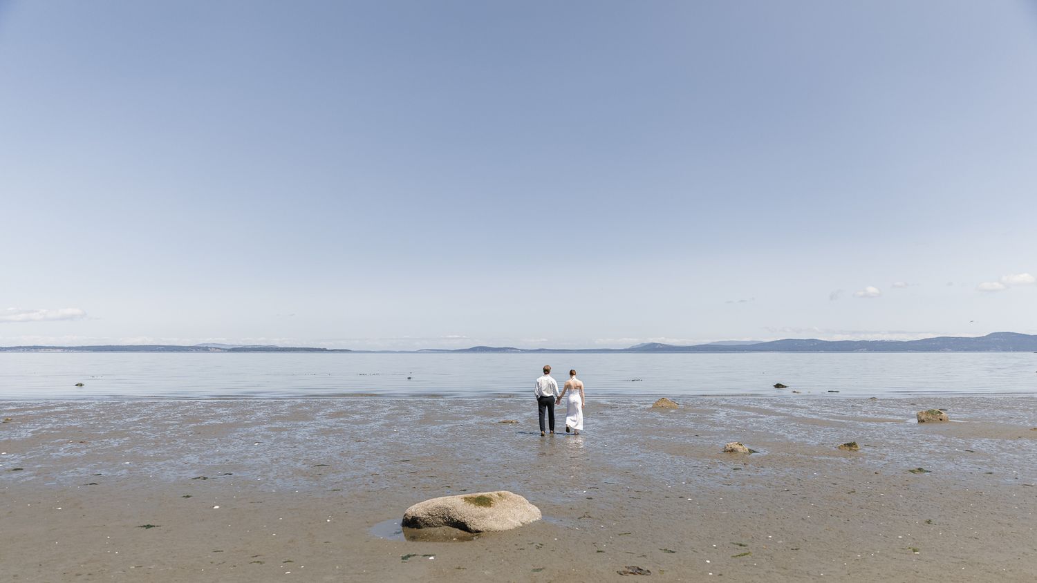 Bride and groom standing together on a serene sandy beach with rocky shoreline and blue sky.