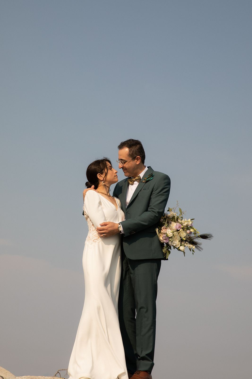 Couple shares romantic moment outdoors, groom holding floral bouquet while bride looks up lovingly.