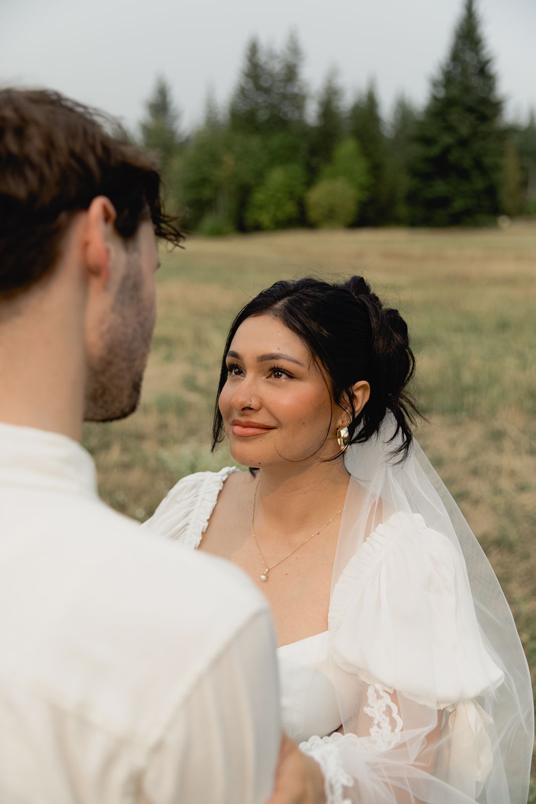 Beautiful dark-haired bride in white wedding dress with veil smiles warmly at groom outdoors.