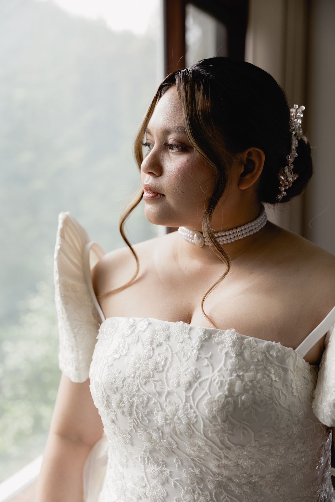 Elegant bride in white lace gown and pearl choker gazes out a window wearing bridal gloves and hair accessories.