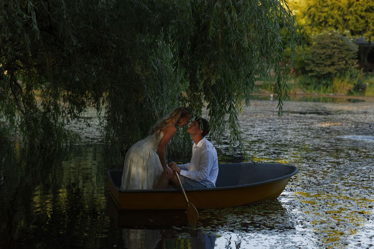 Romantic couple sharing an intimate moment in a wooden rowboat on a tranquil pond beneath a weeping willow tree.