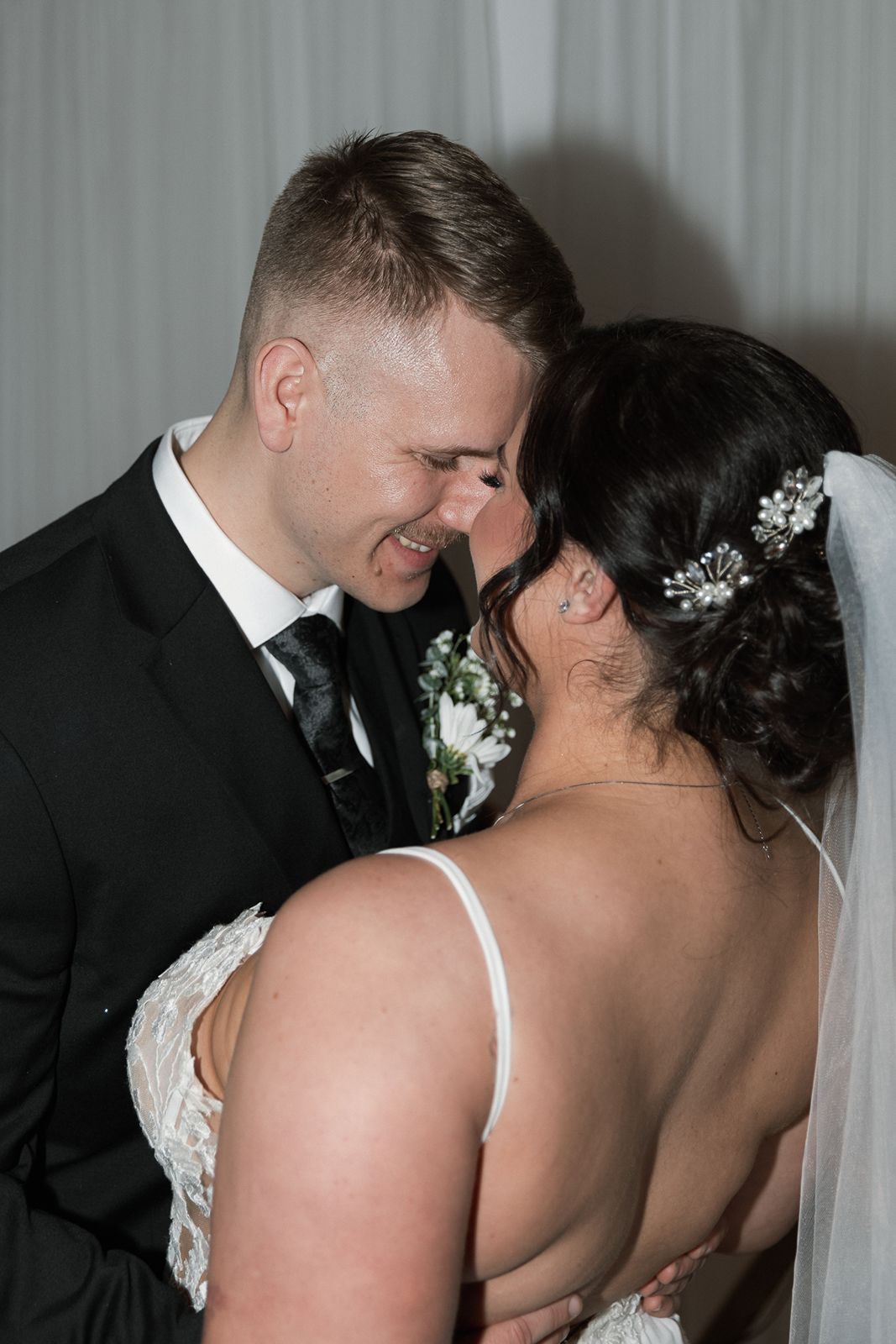 Groom in black suit smiling tenderly at bride with floral hair accessories and lace wedding dress.