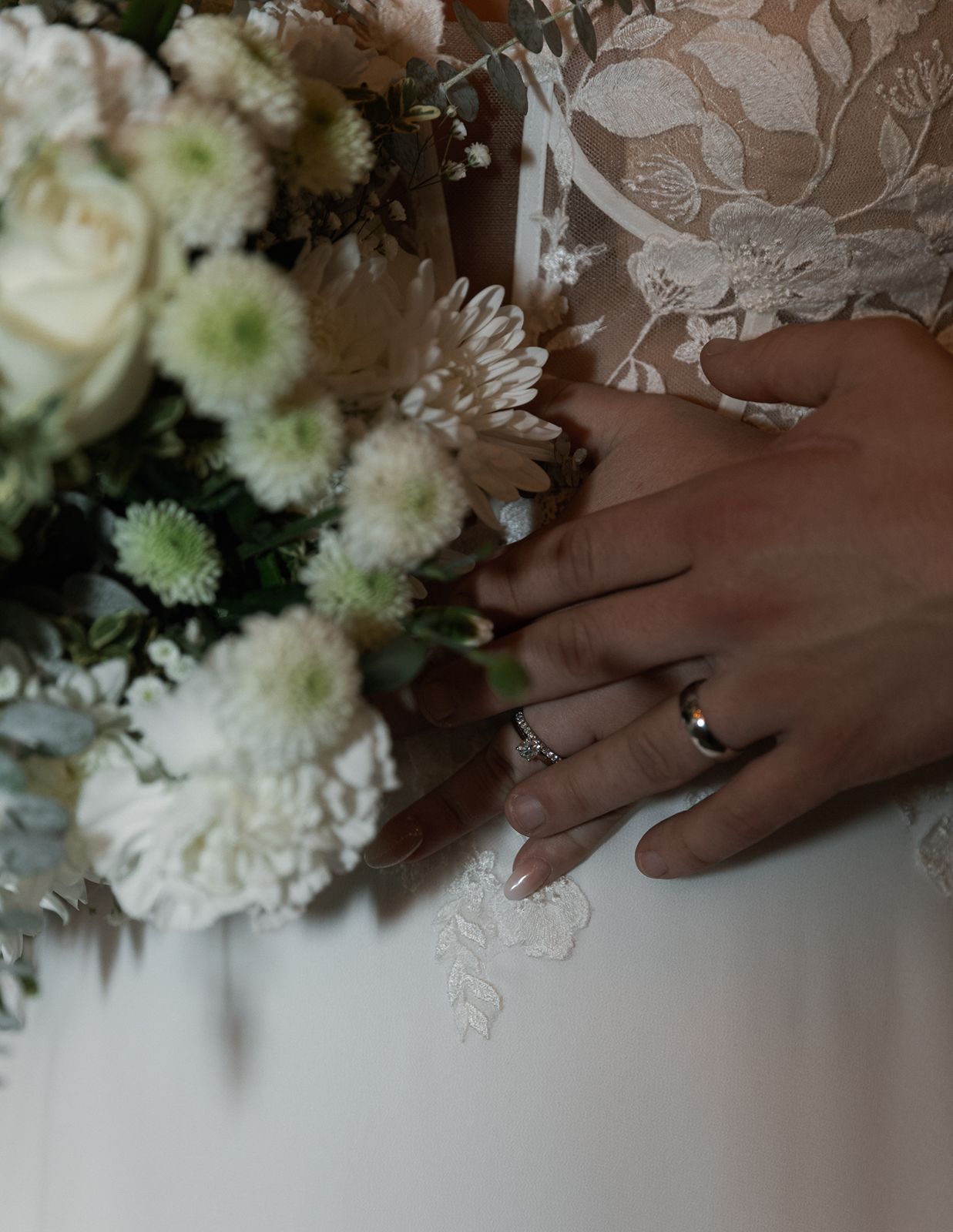 Close-up of newlyweds hands with wedding bands resting on white floral bridal bouquet.