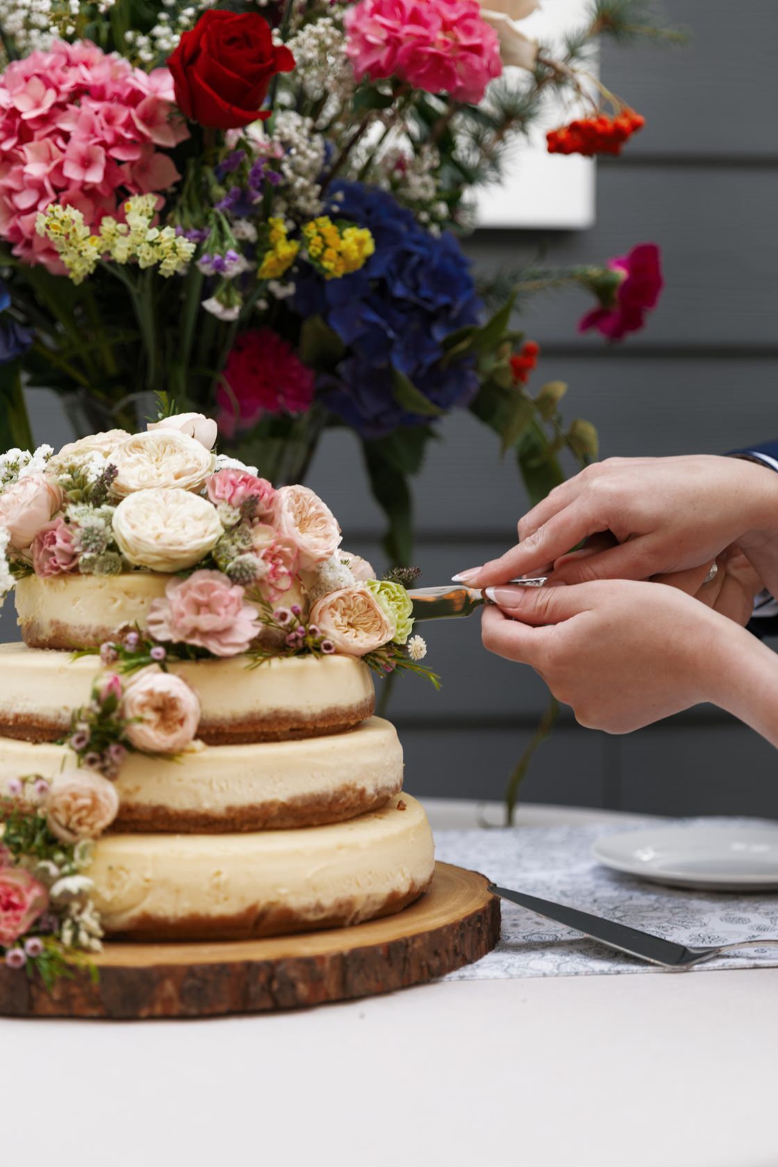 Hands cutting a three-tier naked wedding cake adorned with pink roses, with colorful floral arrangement in background.