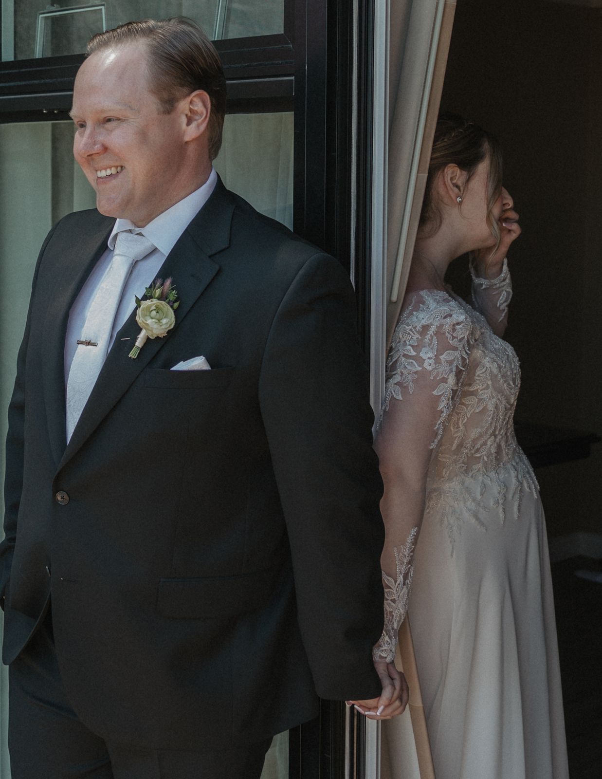 Groom in black suit with boutonniere smiling as bride in lace gown peeks around a curtained doorway.