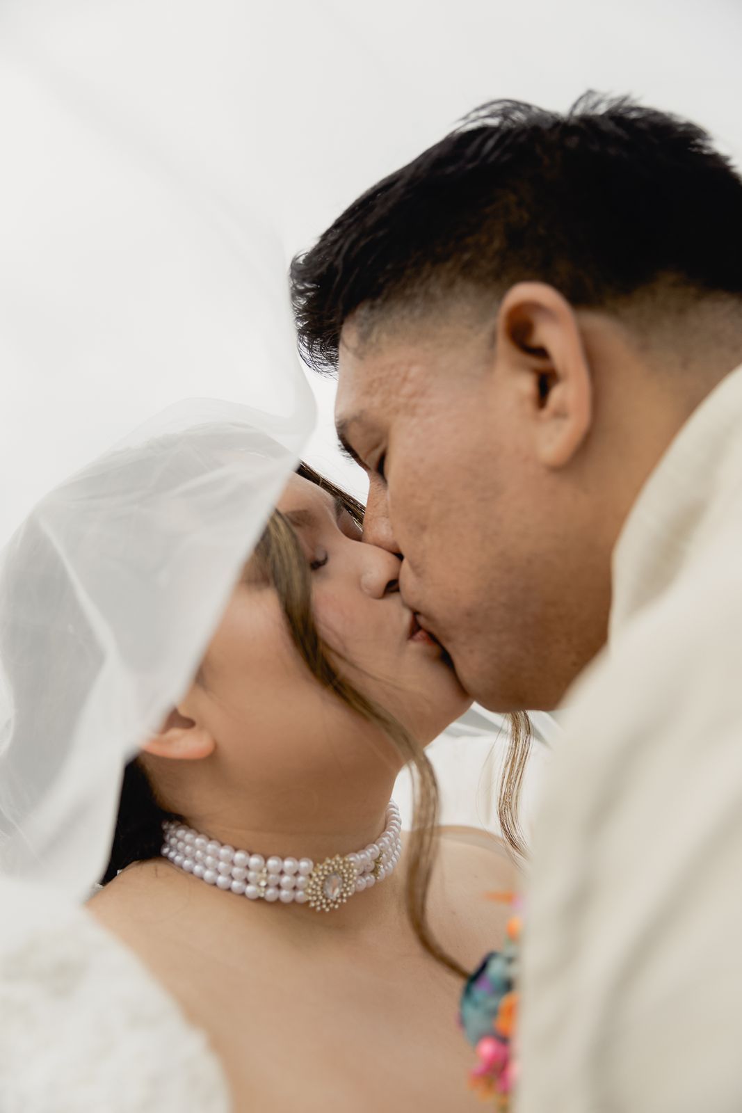 Bride and groom share a romantic kiss beneath a white wedding veil, bride wearing a pearl choker necklace.