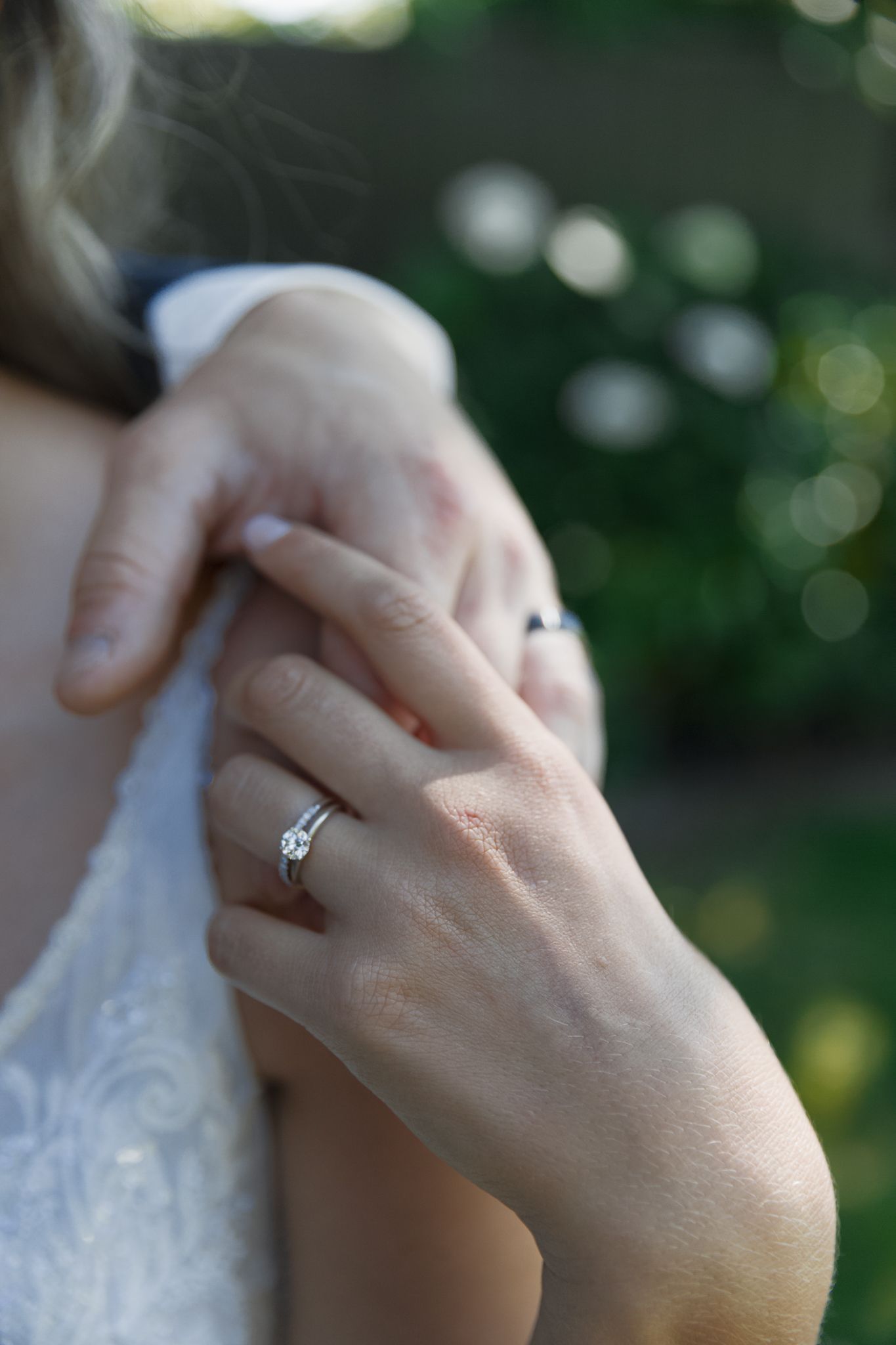 Hands softly holding over a brides shoulder, wedding rings glistening in the warm summer sun.