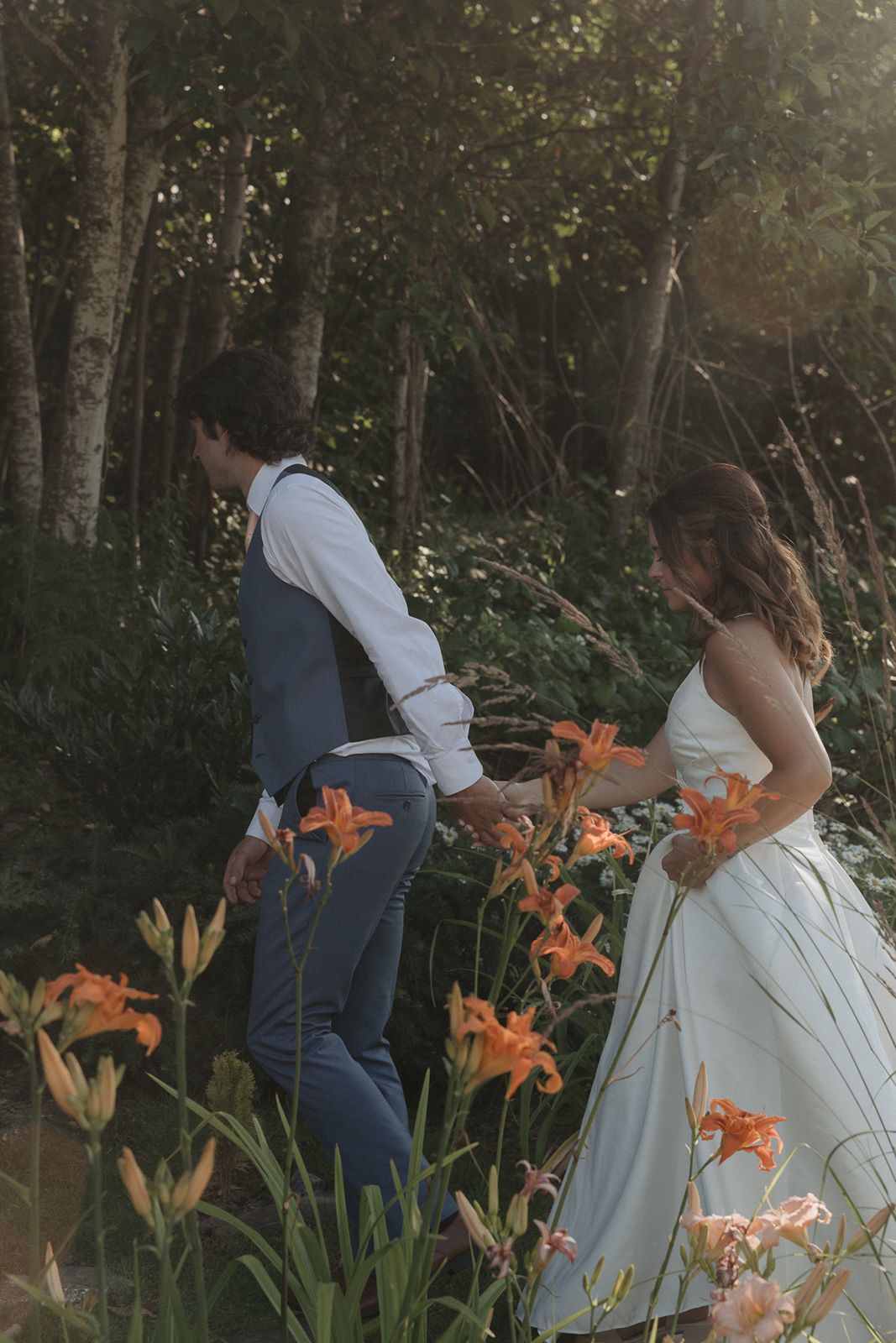 Bride and groom walk hand in hand through a lush garden with vibrant orange lilies during their wedding day.