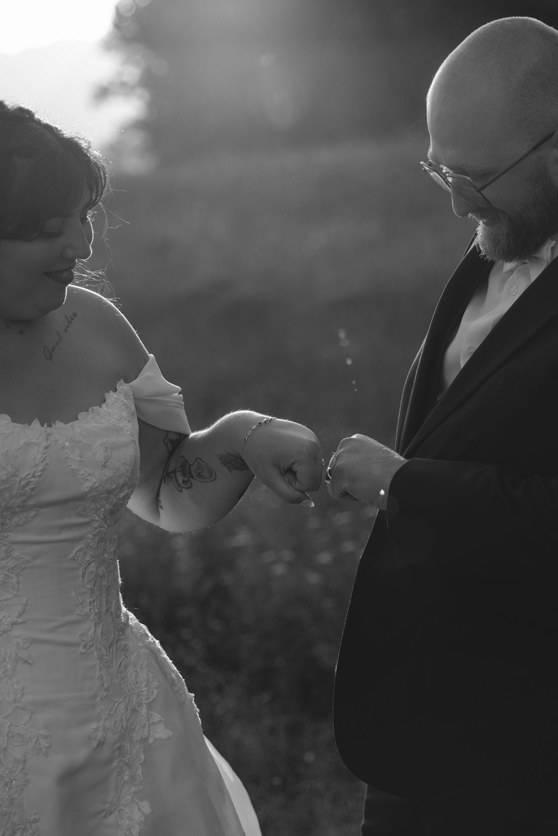 Black and white wedding photo of groom placing ring on bride's finger outdoors in soft backlit sunlight.