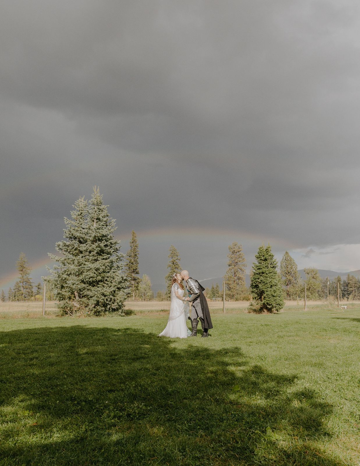 Newlyweds kiss on green lawn with rainbow arching over stormy sky and pine trees.