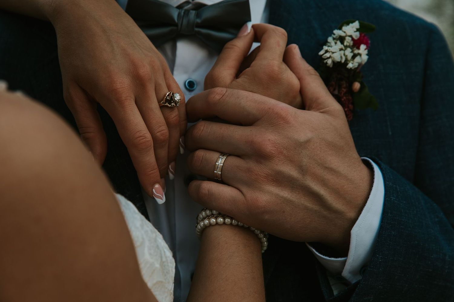 Close-up of bride and groom holding hands, showing wedding rings, pearl bracelet, and boutonniere on wedding day.