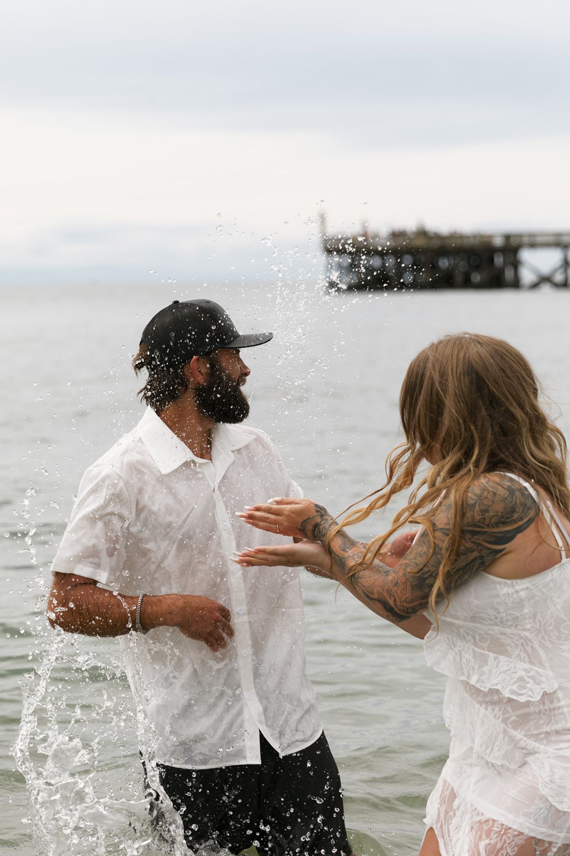 Couple in white outfits splashing water at each other in the ocean near a pier, both with tattoos and wet clothing.