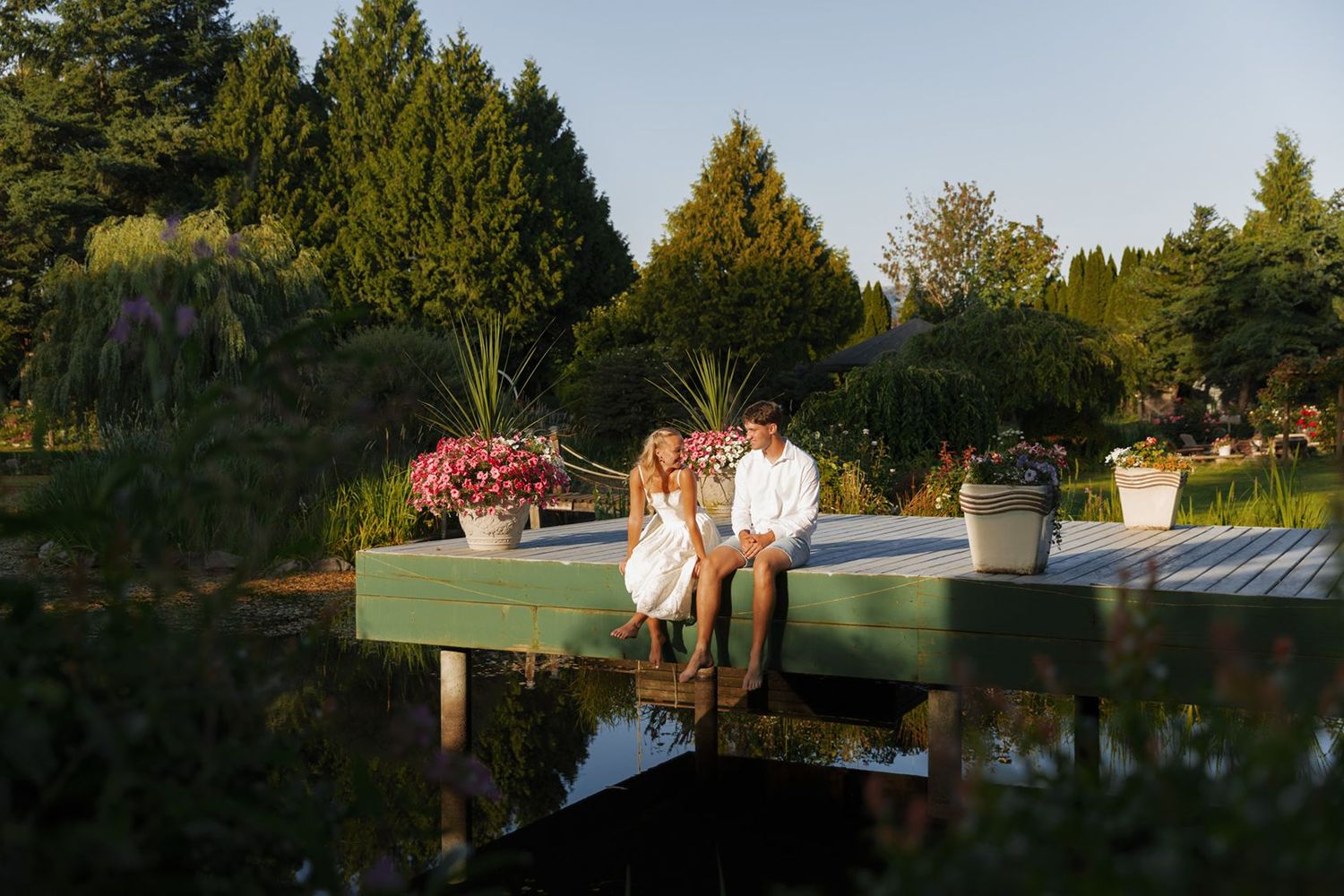 Couple sitting together on a green wooden dock surrounded by flower pots and lush garden trees at golden hour.