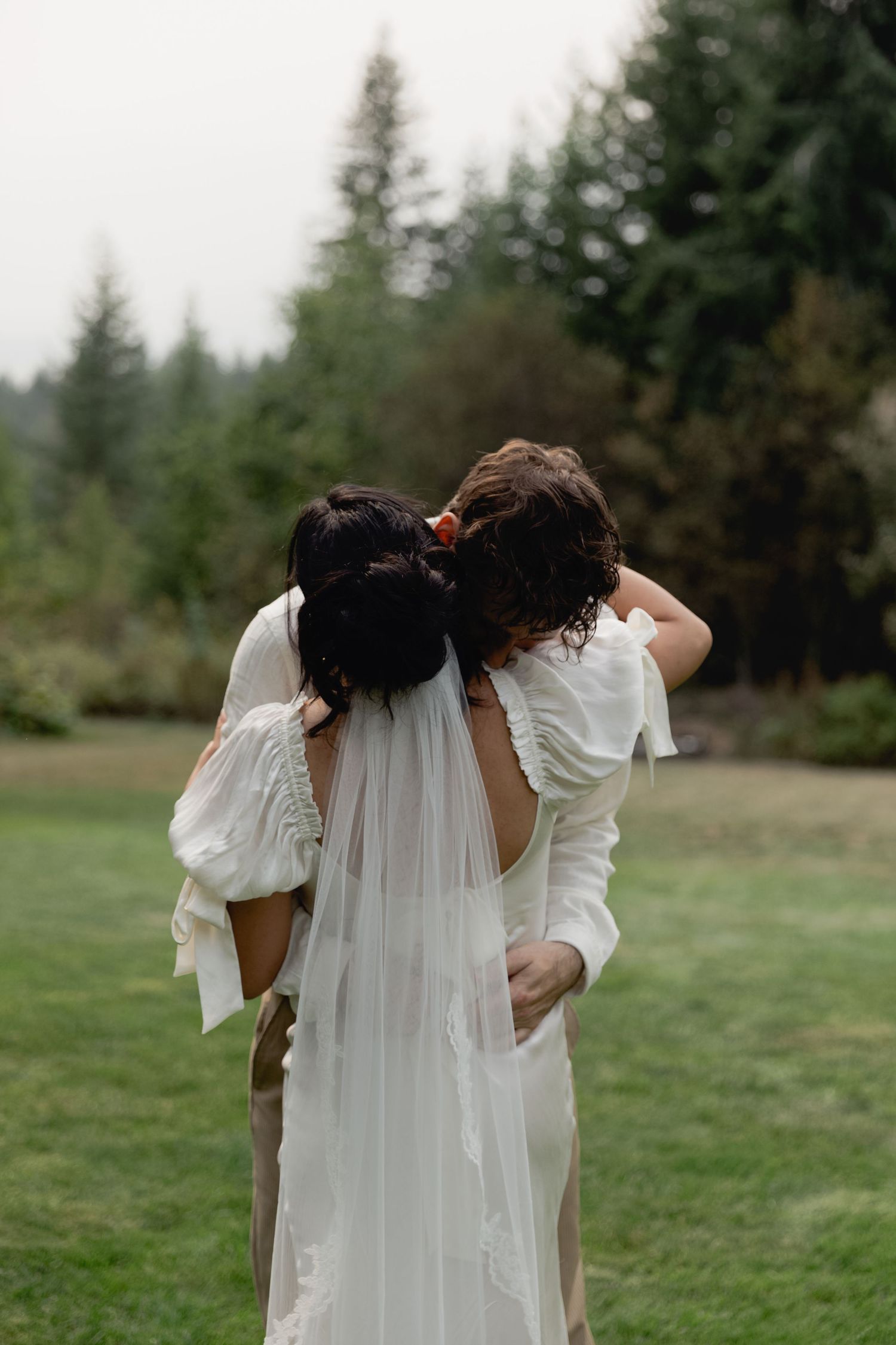 Bride and partner embracing from behind in a forest clearing, bride wearing white gown with veil.