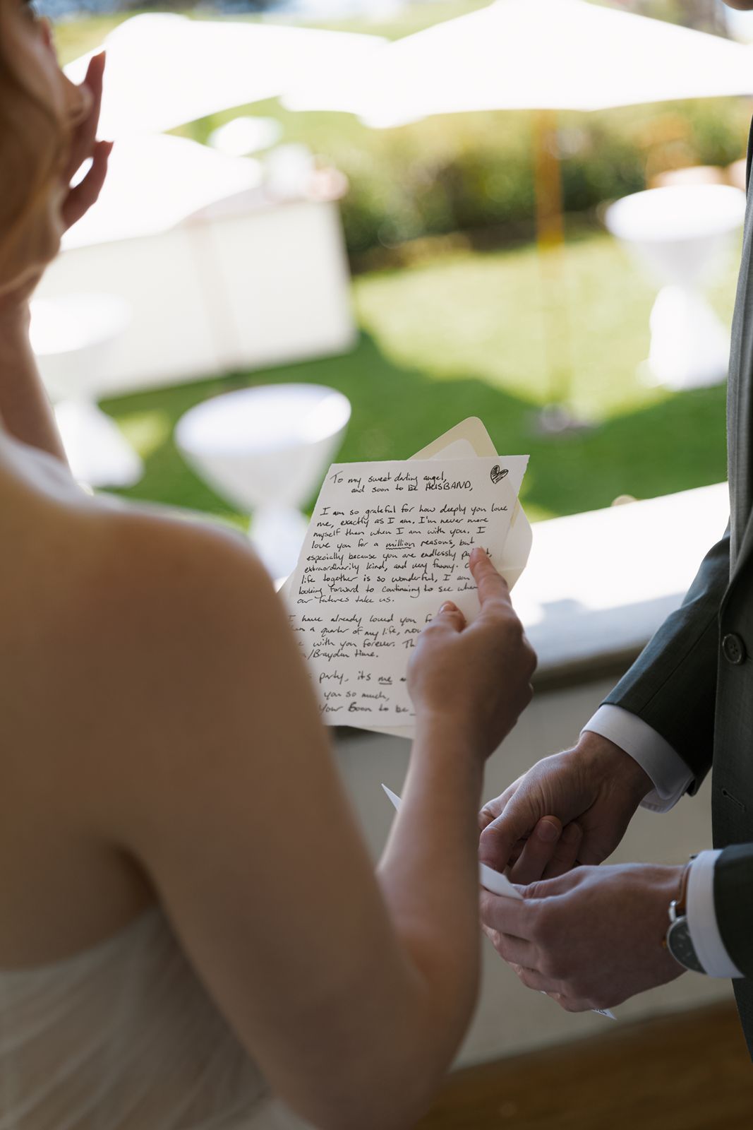 Bride reads handwritten wedding vows while groom holds her hands during outdoor ceremony.
