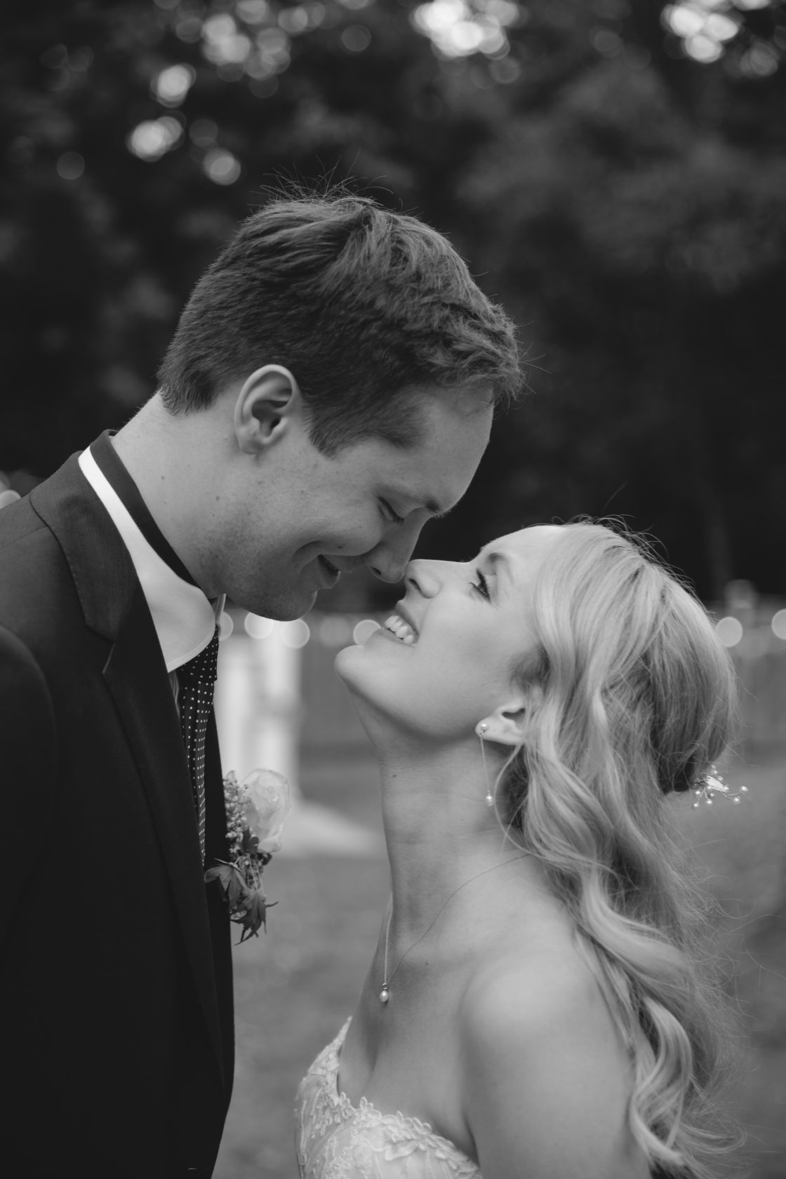 Black and white photo of a smiling bride and groom sharing a romantic moment, nearly kissing outdoors.