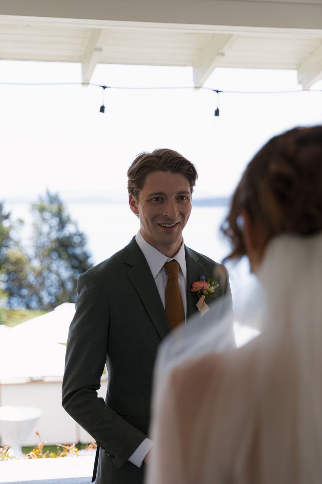 Groom in green suit with orange tie smiling at bride during outdoor wedding ceremony near waterfront.