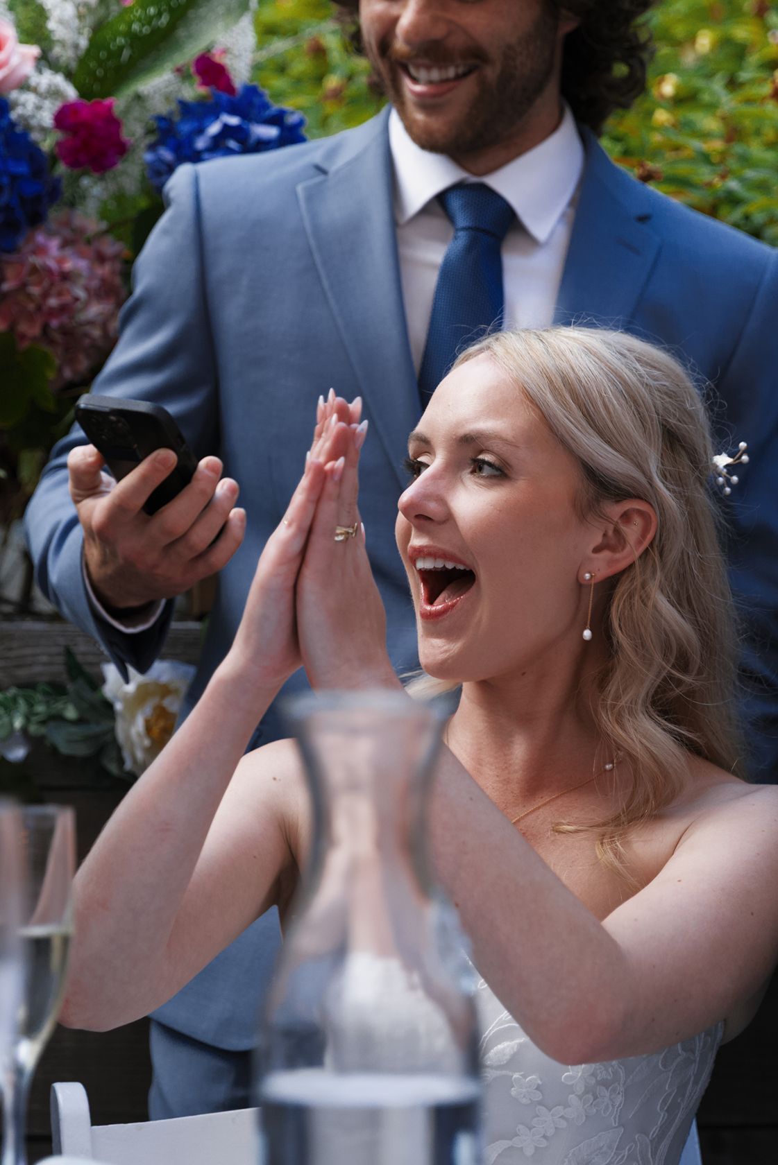 Bride in white dress clapping with joy at wedding reception, groom in blue suit holding phone standing beside her.