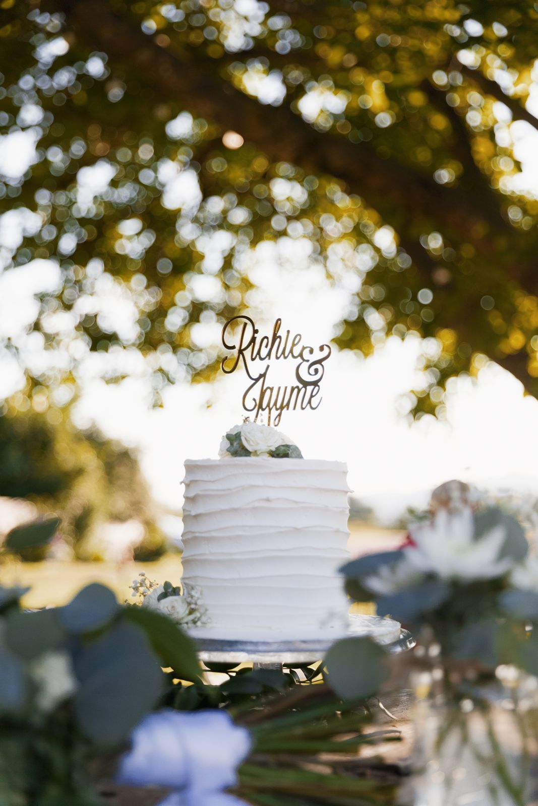 Elegant white wedding cake with Richie and Jayme topper displayed outdoors under a leafy tree.