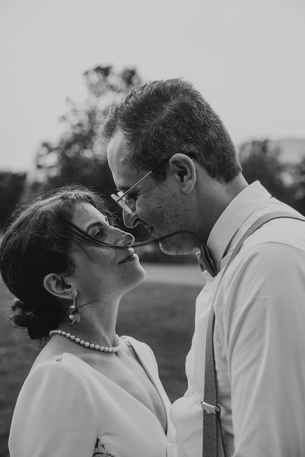 Black and white portrait of bride and groom nearly kissing, bride wearing pearl necklace and glasses.