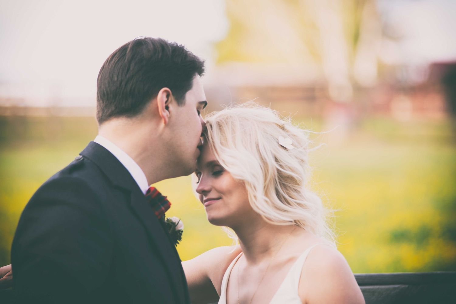 A groom in a dark suit leans in to kiss a smiling blonde bride outdoors at a wedding ceremony.