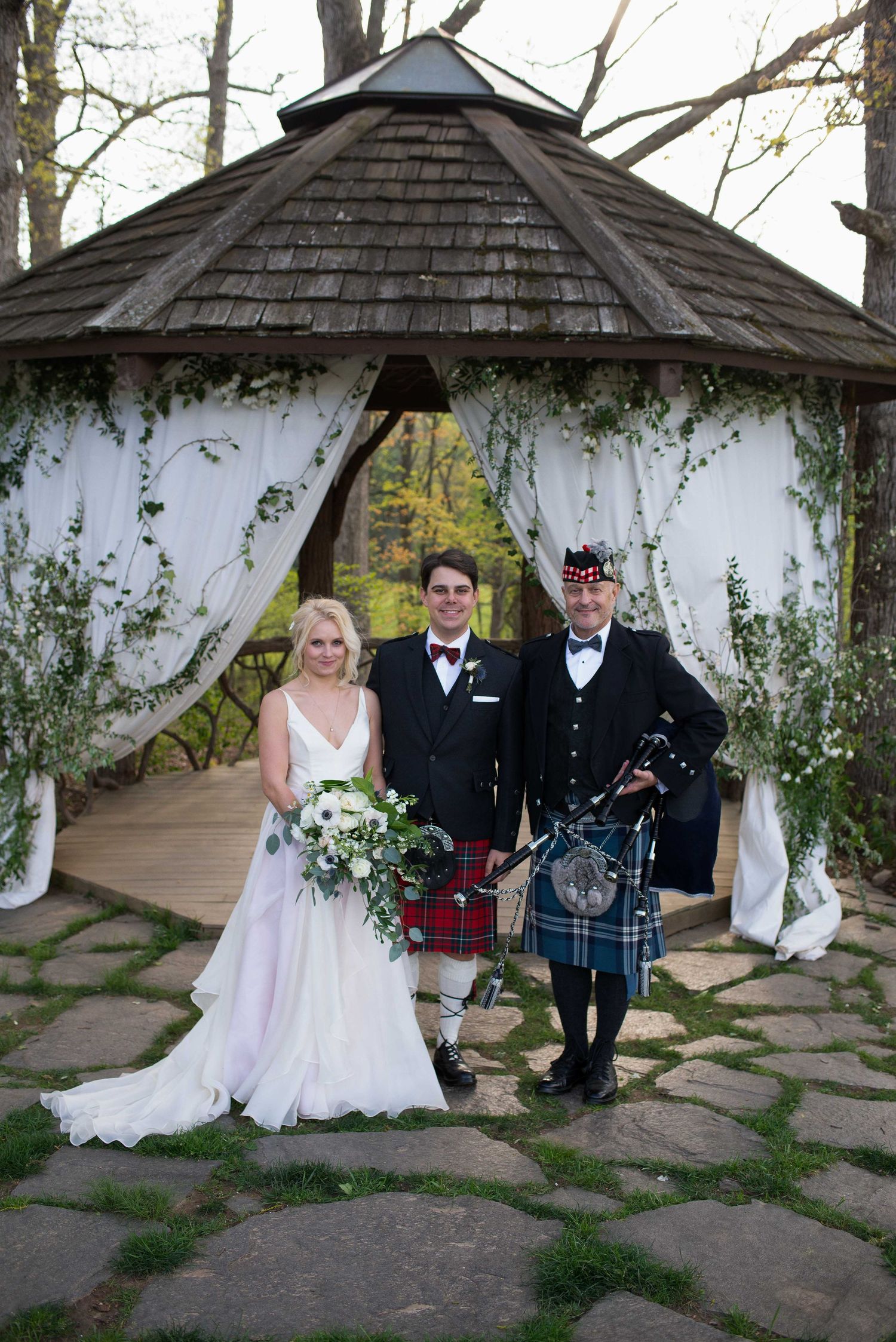 Bride and groom pose with a Scottish bagpiper in a kilt under a decorated gazebo at an outdoor wedding ceremony.