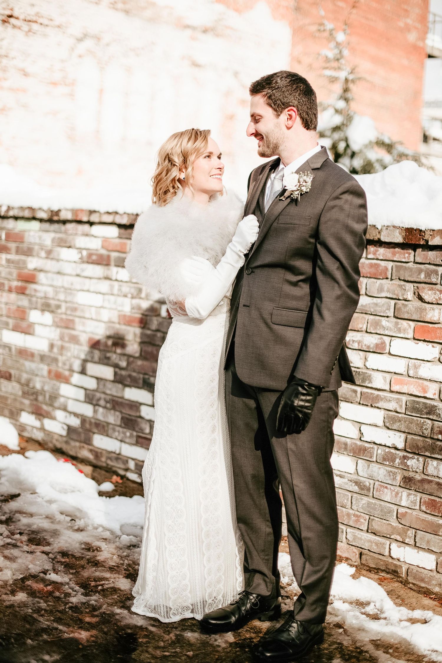 Bride in white lace gown and wrap embraces groom in gray suit beside a snowy brick wall in a winter wedding photo.