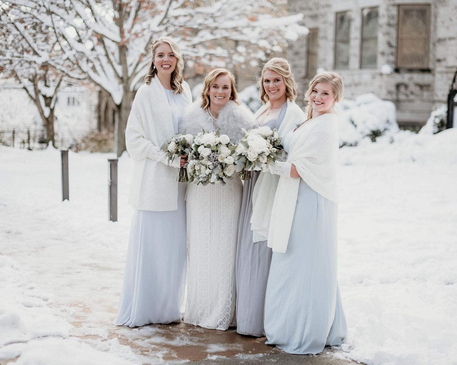 Bride and three bridesmaids in light blue gowns with white wraps holding bouquets outdoors in a snowy winter wedding setting.