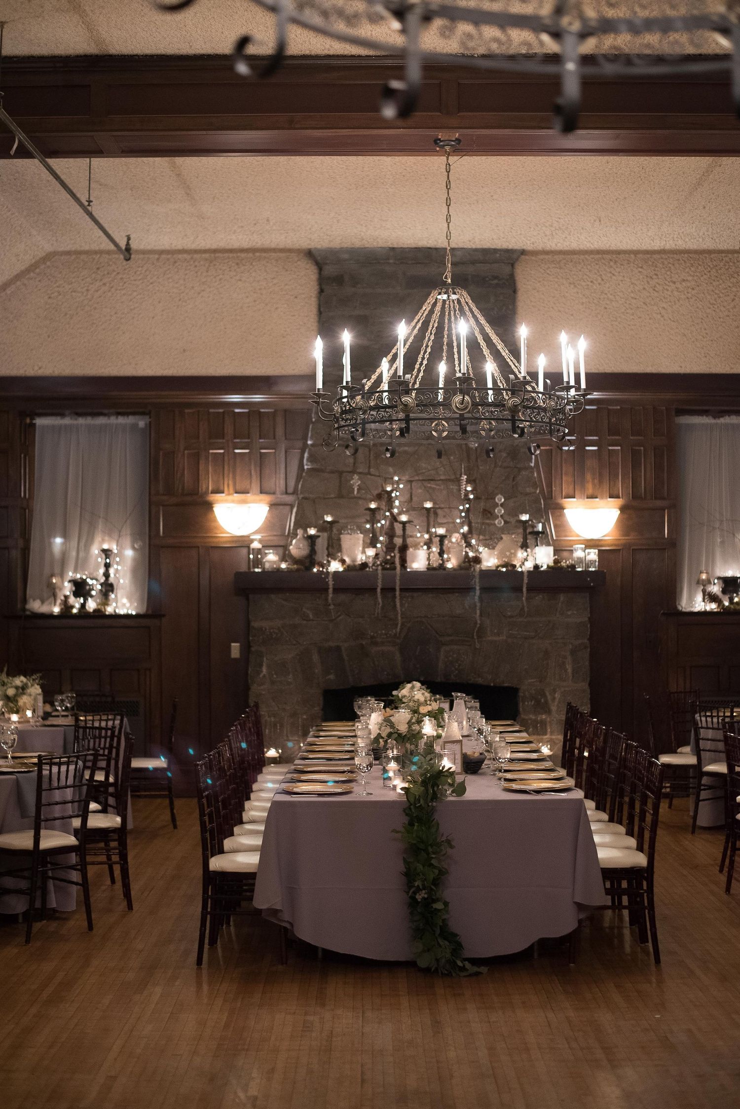 Elegant wedding reception hall with chandelier, stone fireplace, long tables with floral centerpieces, and dark wood paneling.