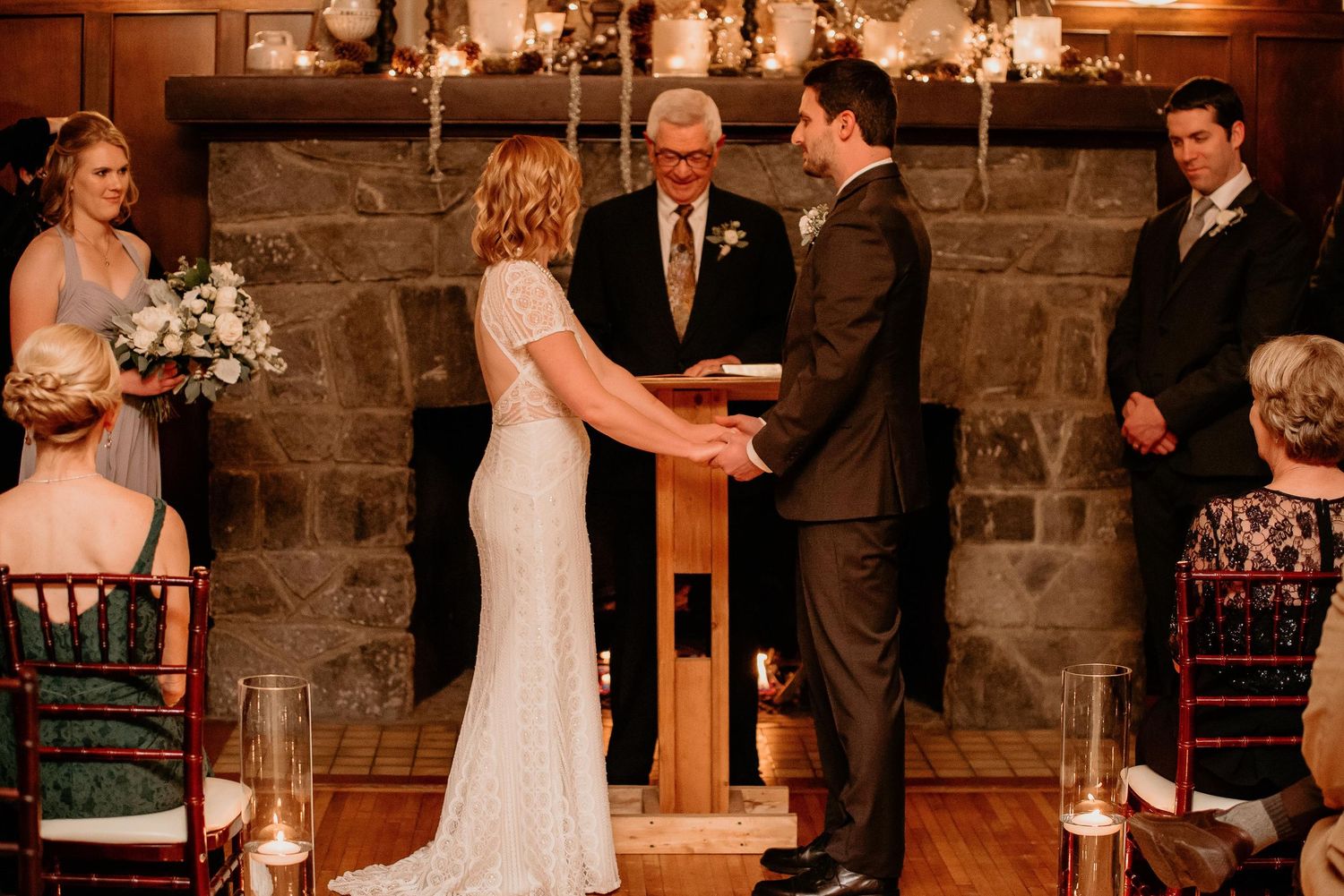 Couple exchanging vows at rustic stone fireplace wedding ceremony with officiant and guests seated nearby.