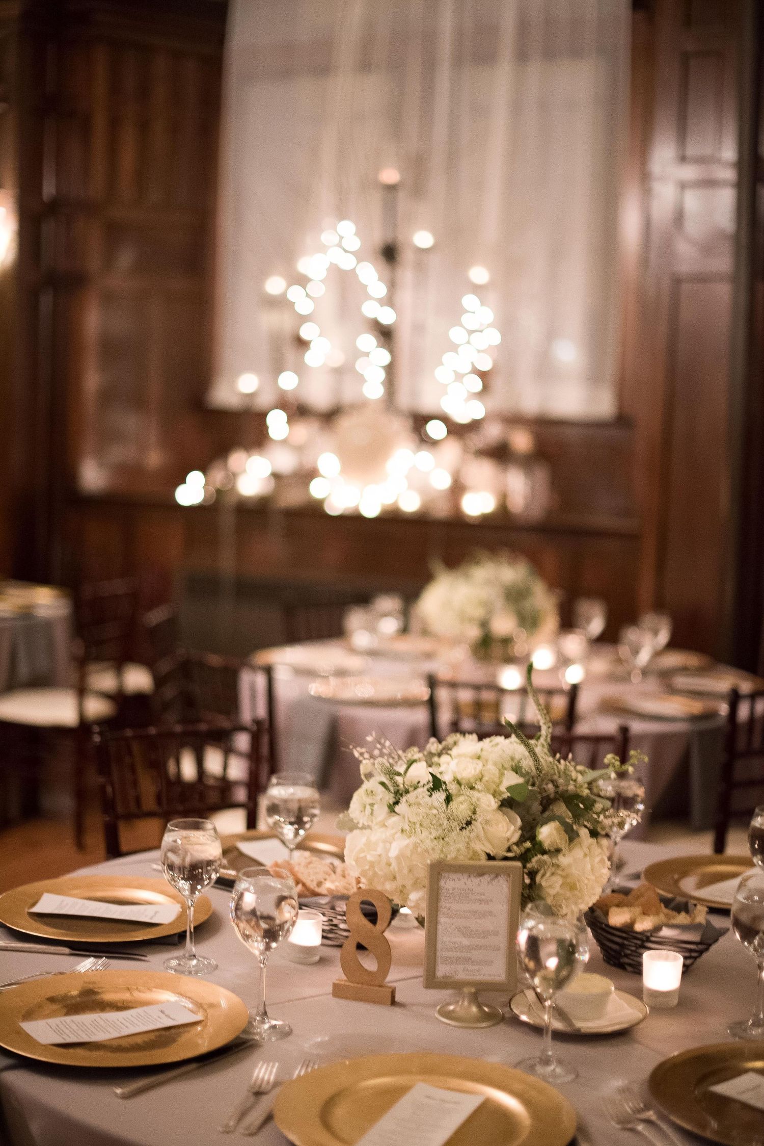 Elegant wedding reception table with gold chargers, white floral centerpiece, crystal glasses, and soft candlelight in a wood-paneled venue.