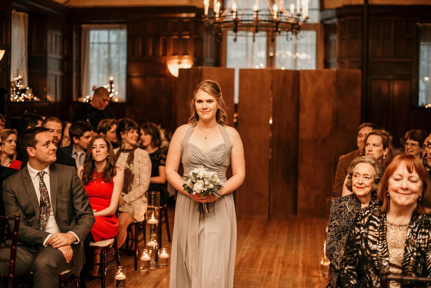 Bridesmaid in gray gown walks down aisle holding white bouquet at elegant wood-paneled wedding ceremony.
