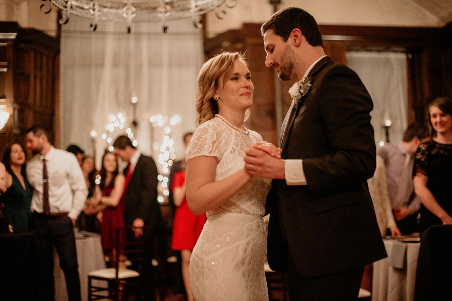 Bride and groom share their first dance at elegant wedding reception with twinkling lights and guests in background.