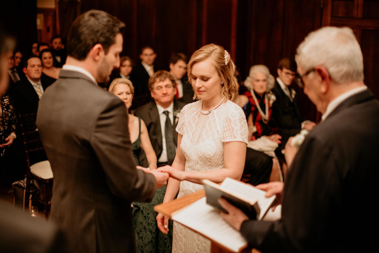 Bride and groom exchange rings during an intimate wedding ceremony while an officiant reads from a book.