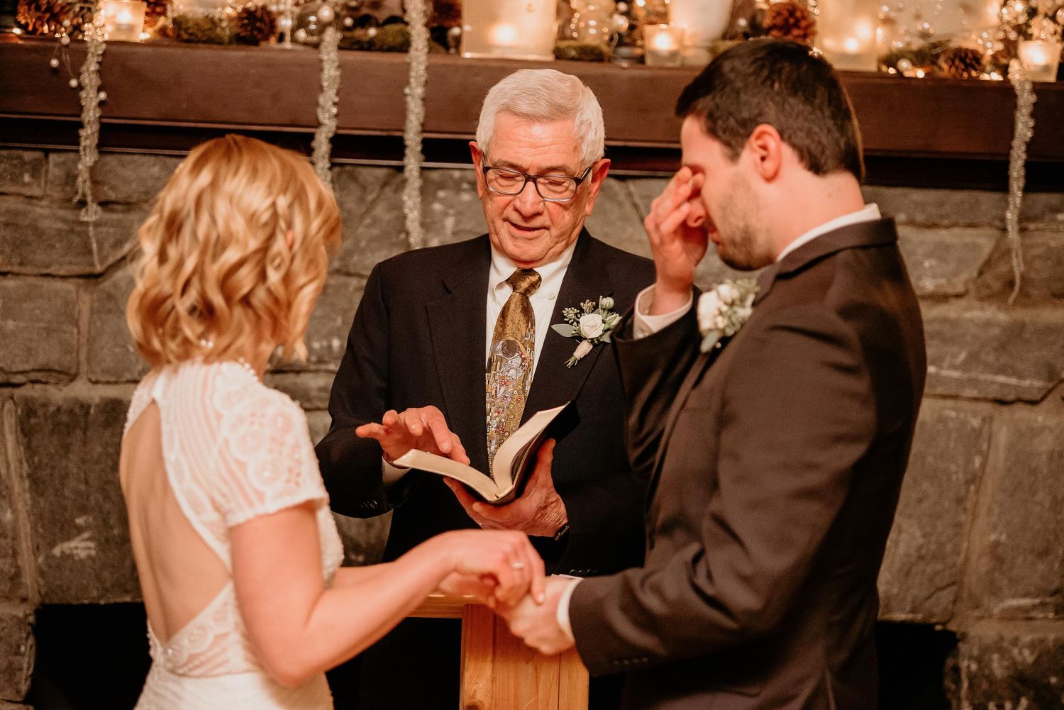 Emotional groom wiping tears during wedding ceremony while officiant reads vows beside fireplace.