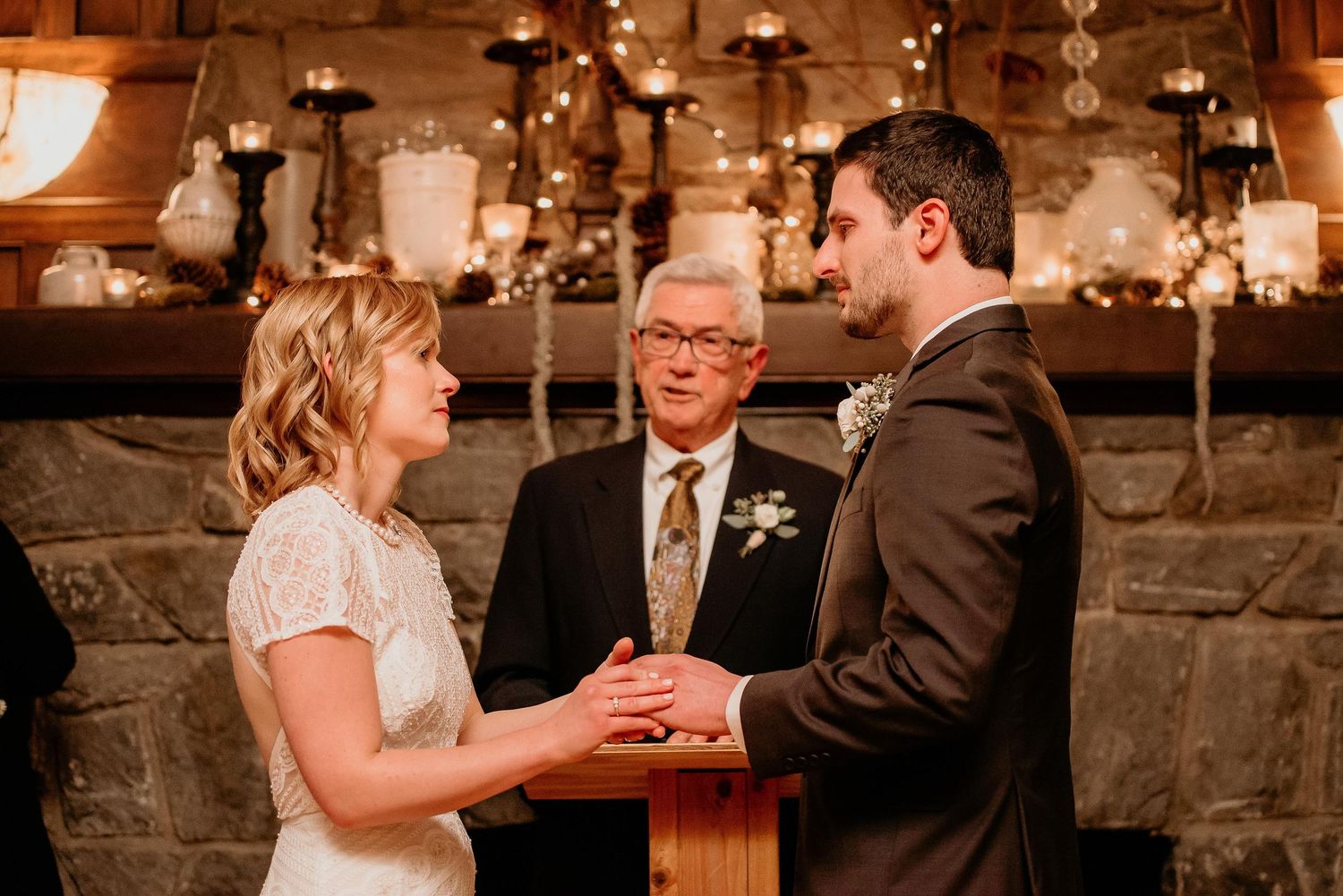 Couple exchanges wedding vows at romantic candlelit ceremony with stone fireplace backdrop and officiant presiding.