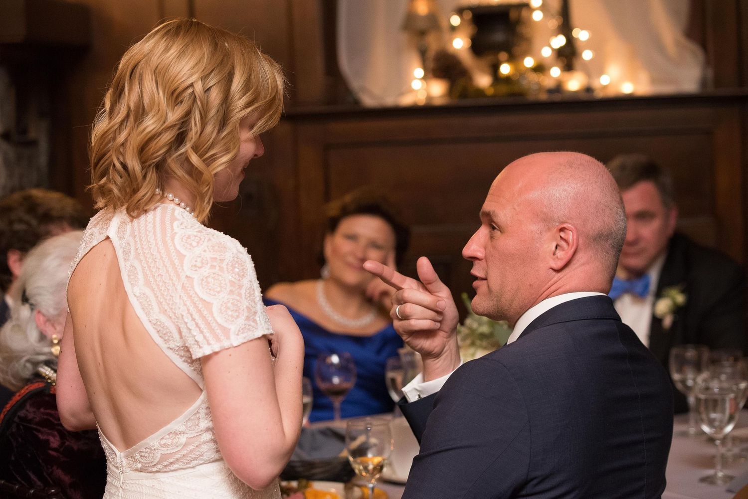 A bald groom gestures while speaking to a bride in a white lace dress at an elegant wedding reception dinner.