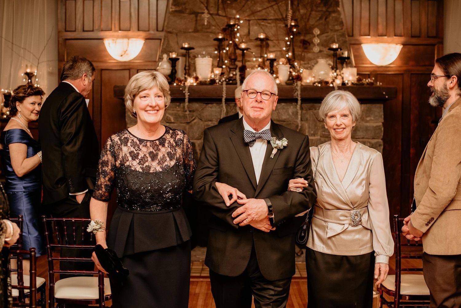 Elegant trio posing at indoor wedding reception near stone fireplace, man in tuxedo flanked by two women in formal attire.