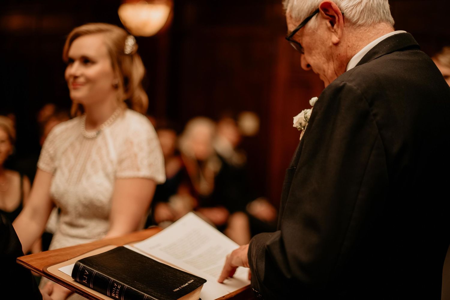Elderly officiant reads from a book at a wedding ceremony podium beside a smiling bride in a white lace dress.