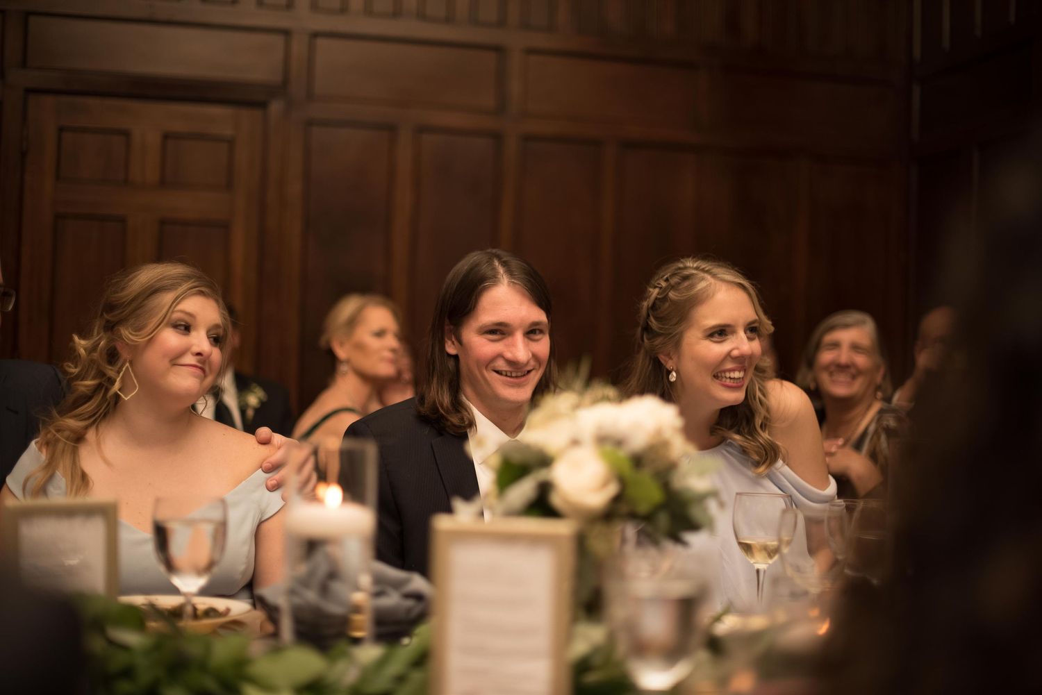 Three guests smiling and laughing together at an elegant wedding reception dinner in a wood-paneled venue.